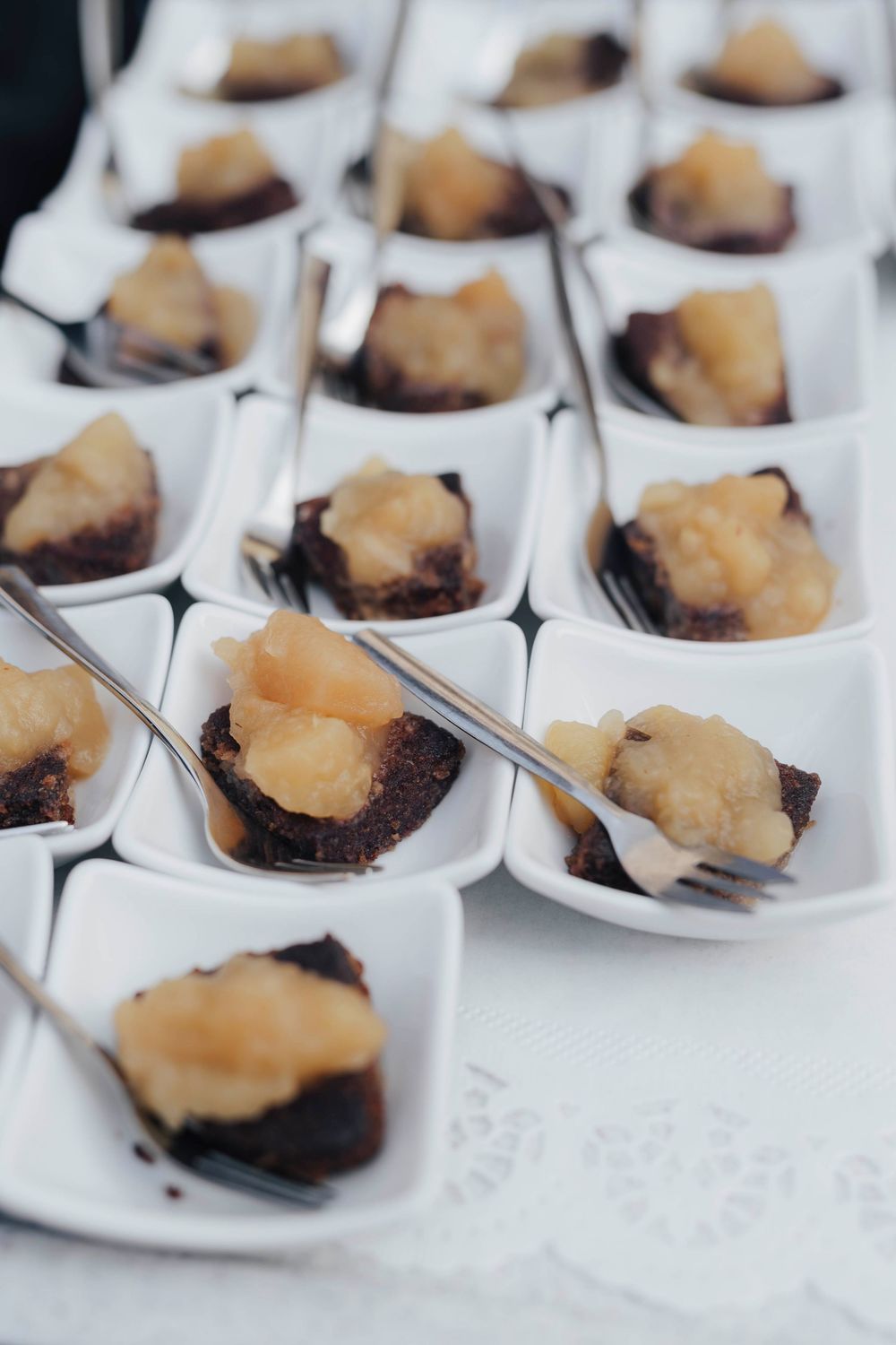White ceramic spoons filled with chocolate brownie bites topped with golden caramelized pear compote arranged on a white tablecloth.