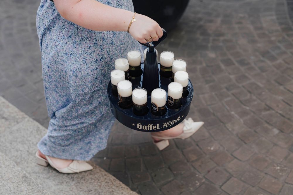 Person carrying a Gaffel Kölsch beer crate with bottles outdoors on cobblestone pavement.