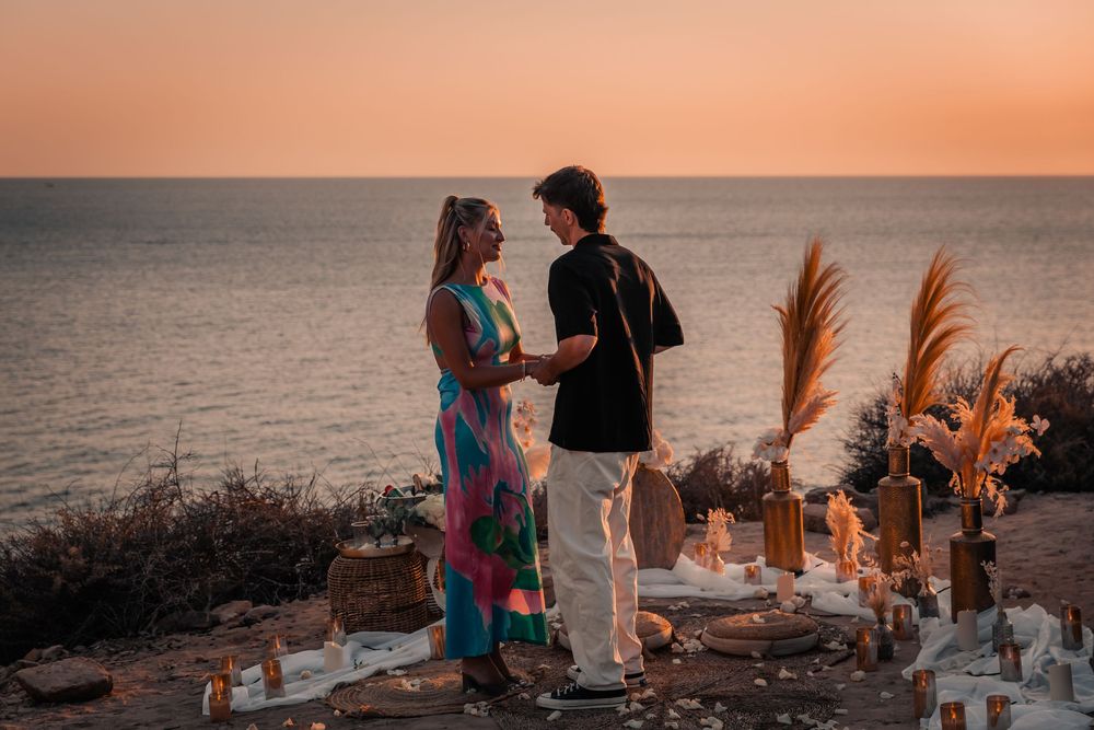 Wide shot of a newly engaged couple standing together next to a bohemian cliffside arrangement during their Mallorca sunset proposal photosh