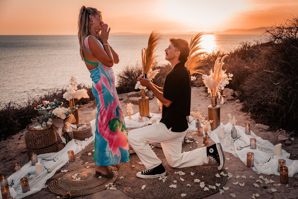 A man down on one knee proposing to a shocked woman during a beautiful sunset surprise proposal in Mallorca.