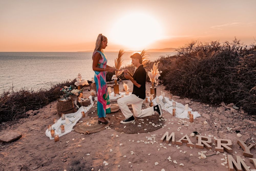 Wide shot of a couple at sunset next to a bohemian cliffside setup celebrating a romantic surprise proposal in Mallorca.
