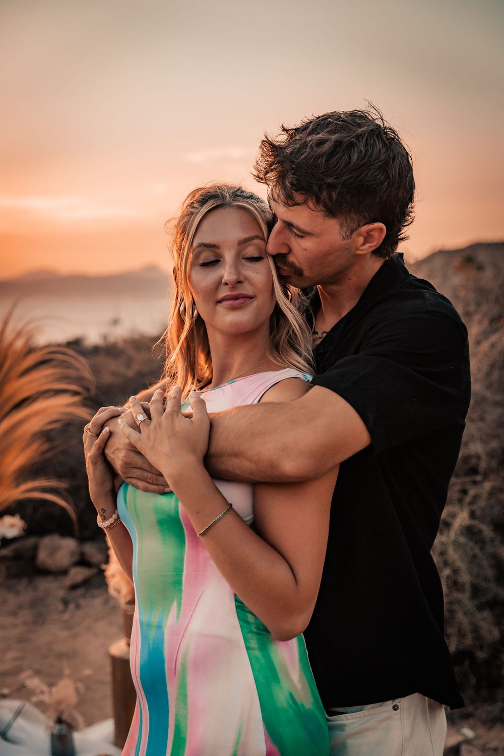 A man holding a woman from behind in an affectionate embrace during a sunset surprise proposal photoshoot on the Mallorca coast.