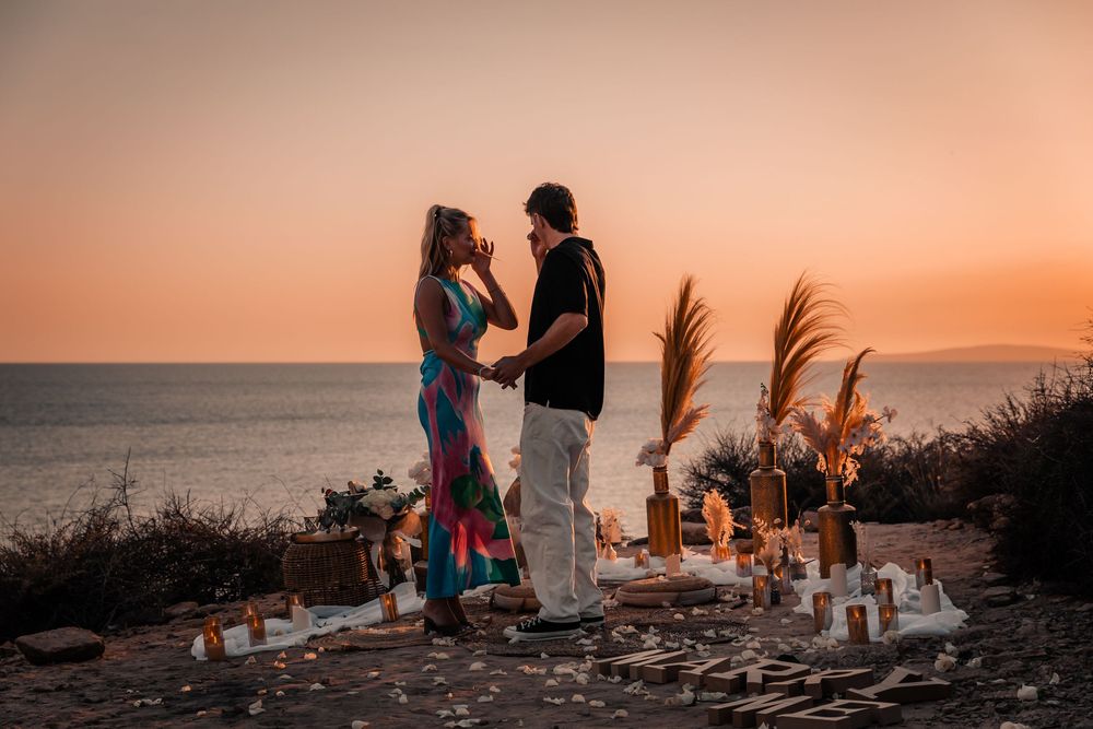 A romantic moment where a woman reacts with emotion during a sunset surprise proposal setup on the Mallorca coast.