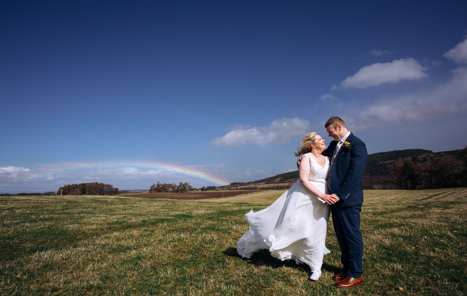 Bride and groom share a romantic moment in a green field with a rainbow and blue sky backdrop on their wedding day.