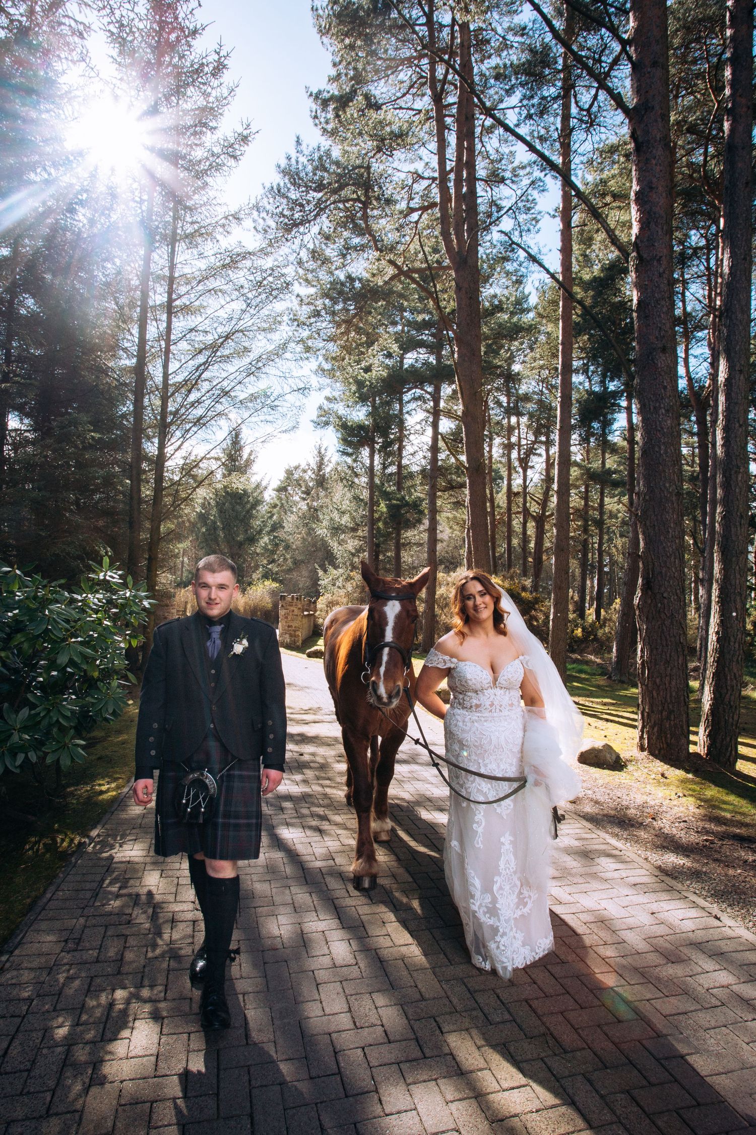 Bride in white gown and groom in Scottish kilt walk with a brown horse along a sunlit forest path.