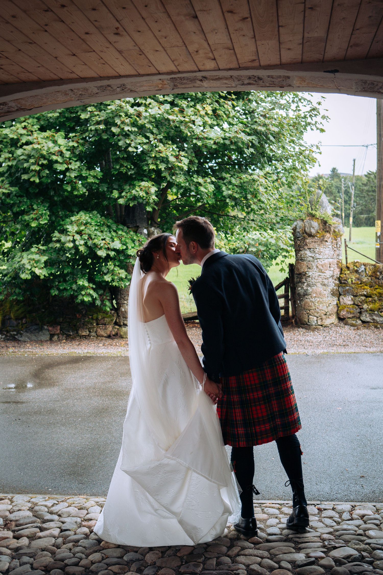 Bride and groom kissing under a wooden arch, groom wearing a red tartan kilt, lush green trees in the background.