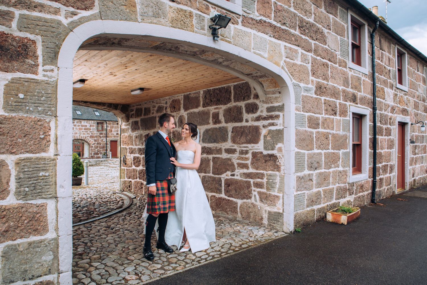 Bride and groom stand under stone archway at Scottish wedding venue, groom wearing traditional tartan kilt.