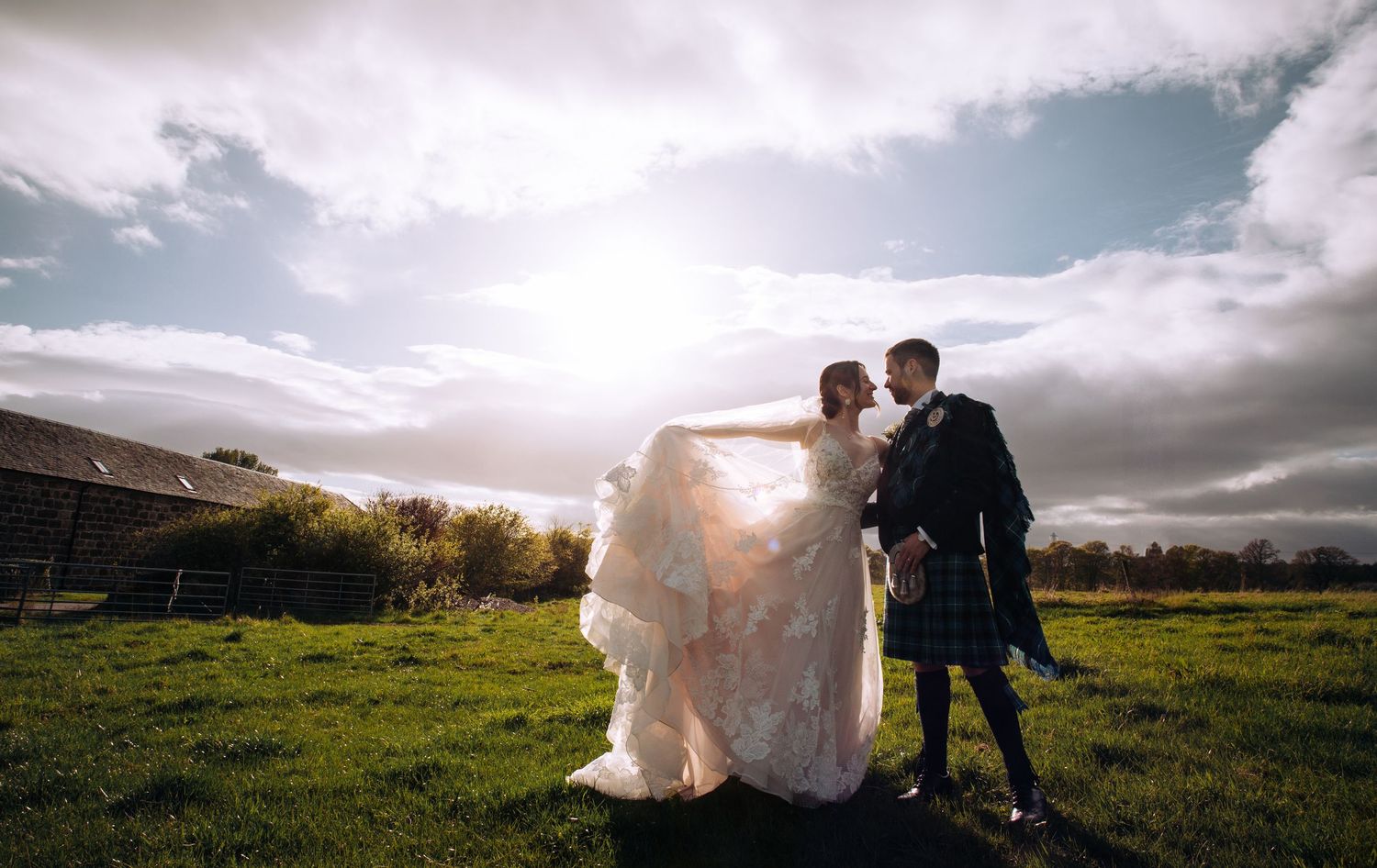 Bride in flowing white gown and groom in tartan kilt embrace on green field with dramatic cloudy sky backdrop.