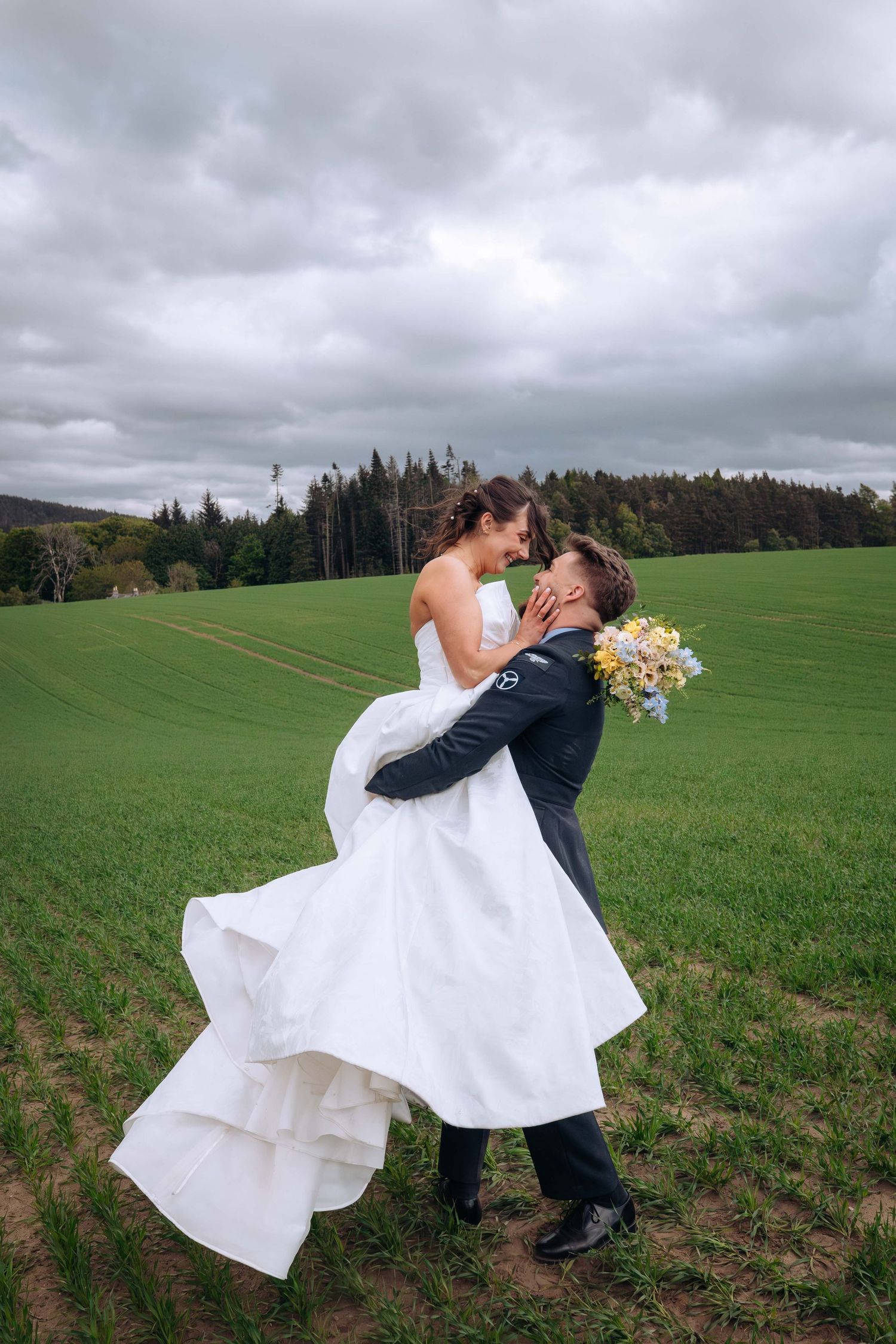 Groom lifts bride in white gown on green field, sharing romantic moment with bouquet, trees and cloudy sky behind them.