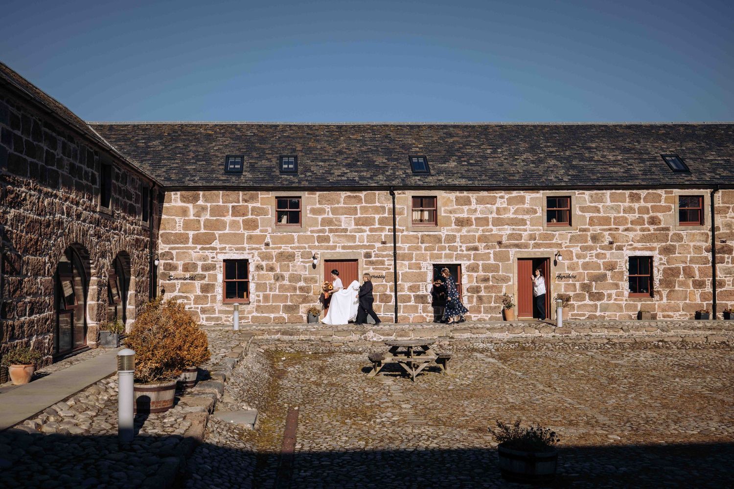 Wedding couple poses in courtyard of rustic stone barn venue under clear blue sky.