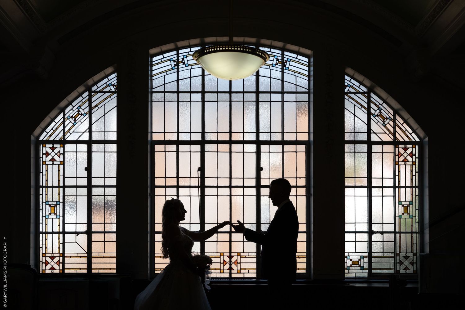 Silhouette of a couple holding hands in front of an ornate stained glass arched window.