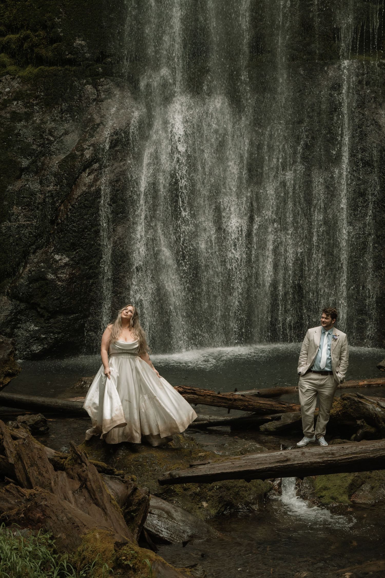 Bride and groom stand before a stunning waterfall on rustic wooden logs in a lush forest wedding setting.