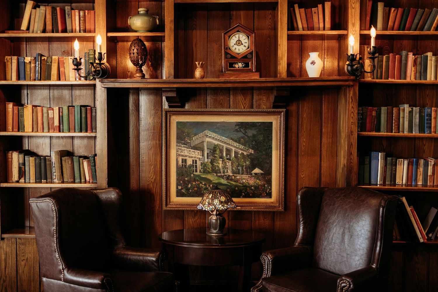 Cozy vintage library with leather chairs, wooden bookshelves, candles, and a framed painting above a small round table.