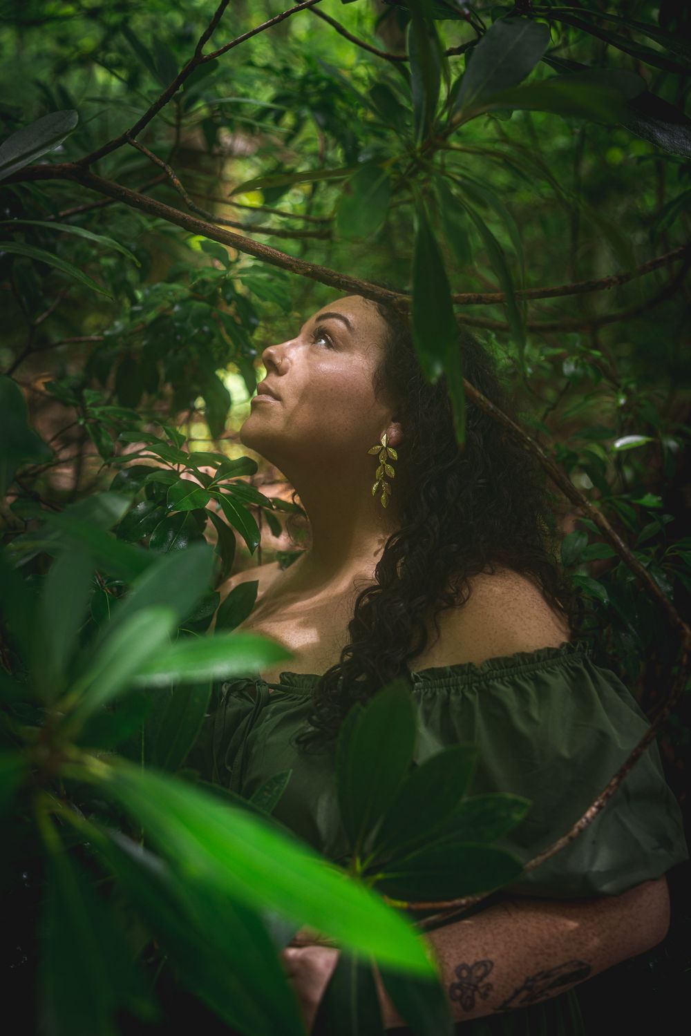 Woman in green off-shoulder top gazes upward surrounded by lush tropical foliage in a moody forest setting.
