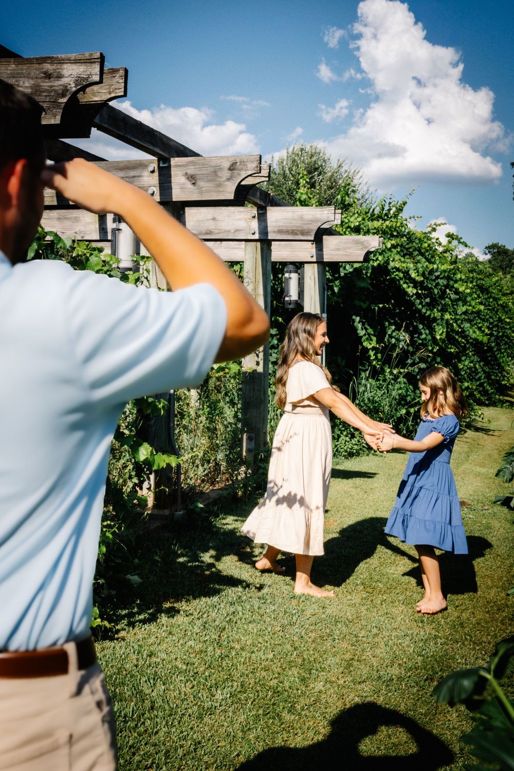 A man photographs a woman and young girl holding hands in a lush garden under a wooden pergola on a sunny day.