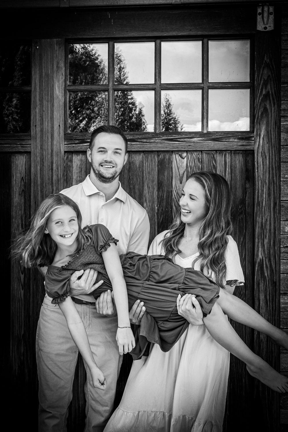 Black and white family portrait of smiling parents holding a child in front of a rustic wooden door.