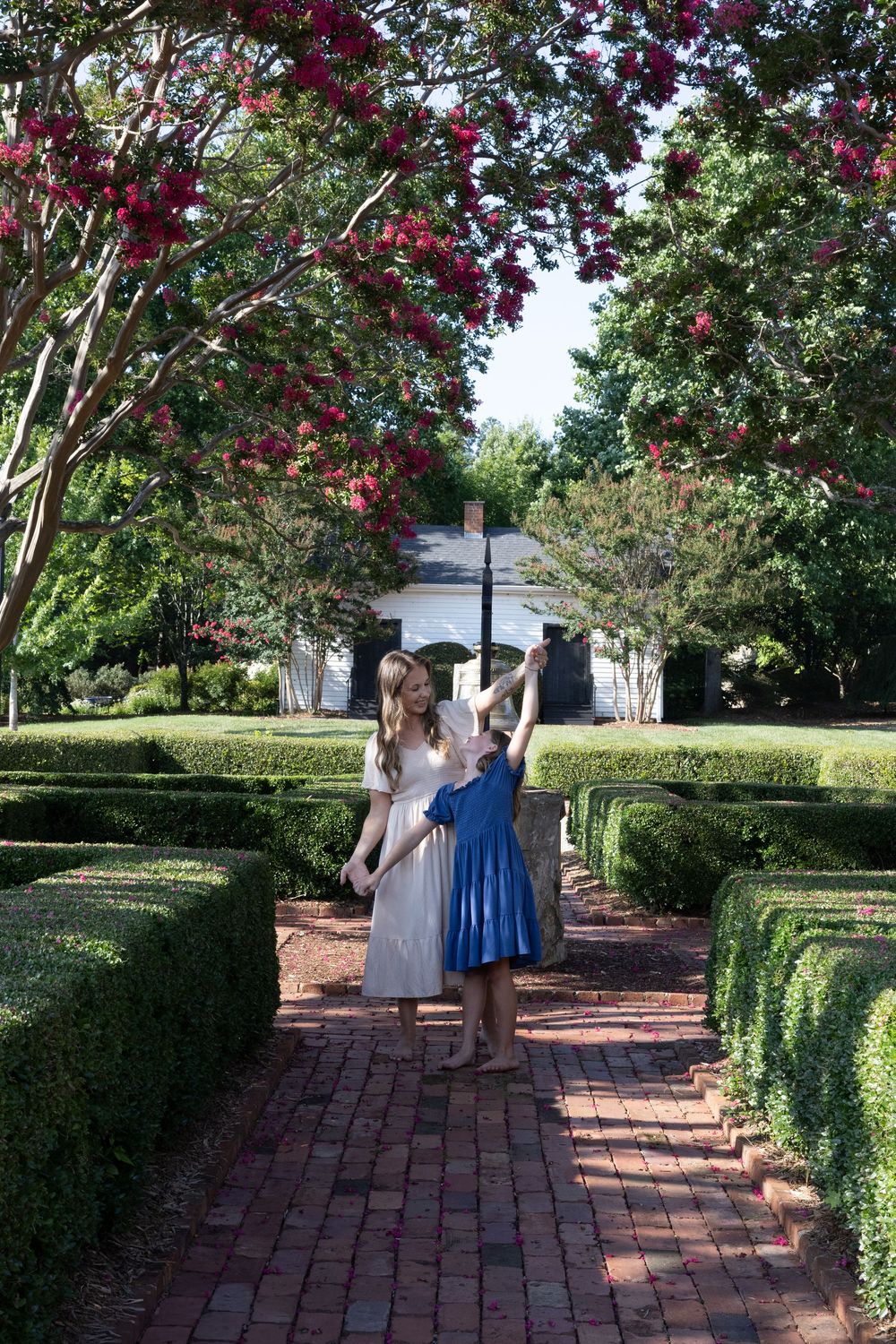 Two girls in dresses walk along a brick path through manicured hedges beneath blooming red crepe myrtle trees.