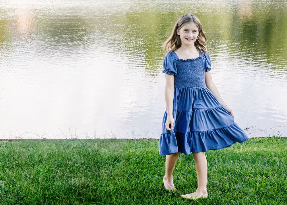 A young girl in a blue tiered dress stands barefoot on green grass near a serene lake, smiling gracefully.