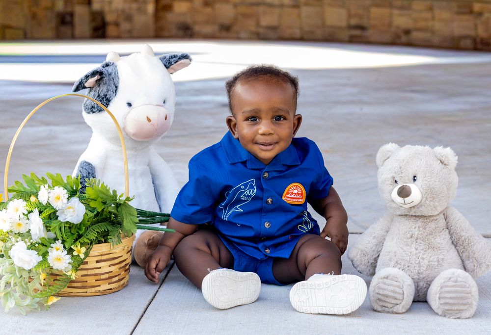 Adorable baby boy in blue outfit sitting between a stuffed cow, flower basket, and teddy bear outdoors.
