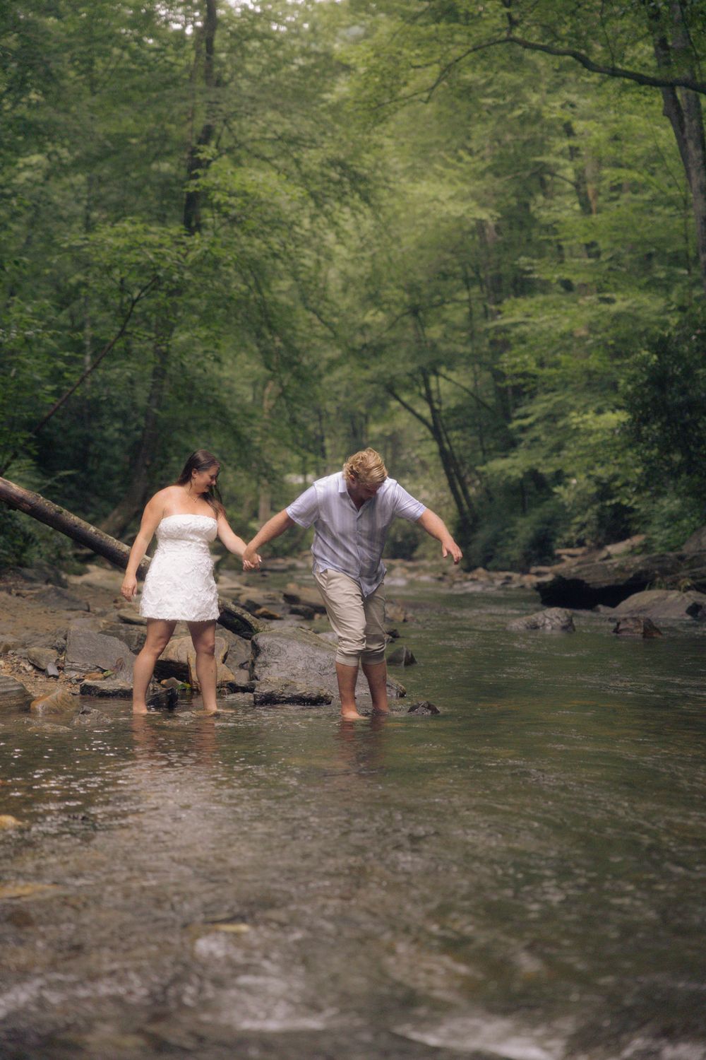 Couple holding hands wading through a shallow forest creek, woman in white dress and man in casual attire.