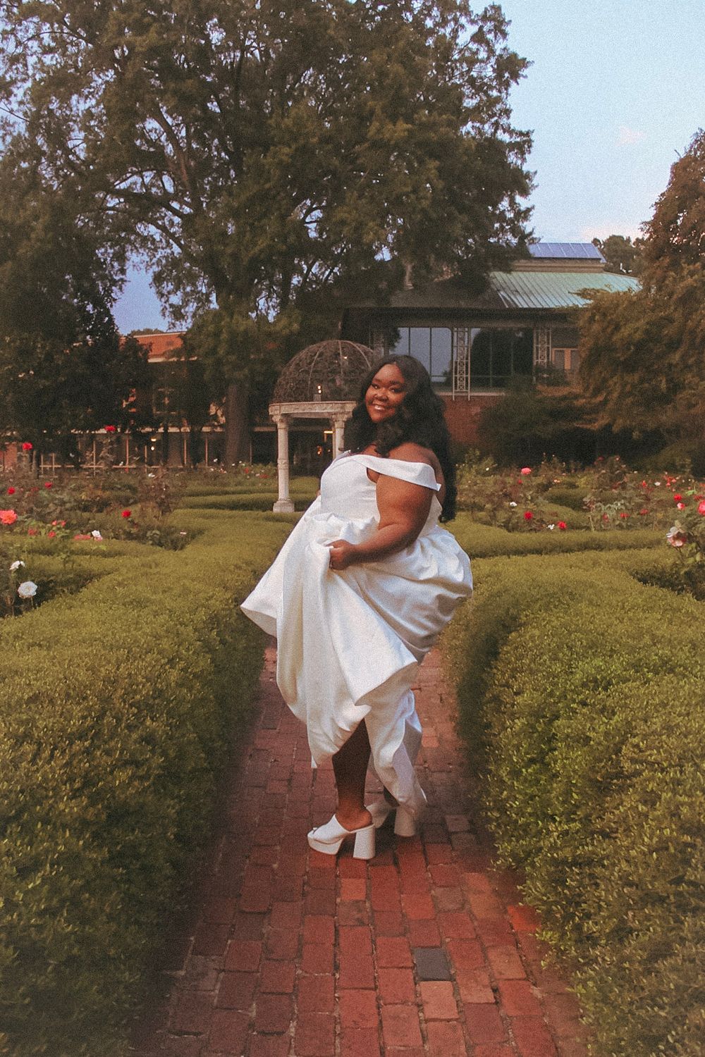 Woman in white off-shoulder gown poses on brick path in formal garden with hedges and roses.