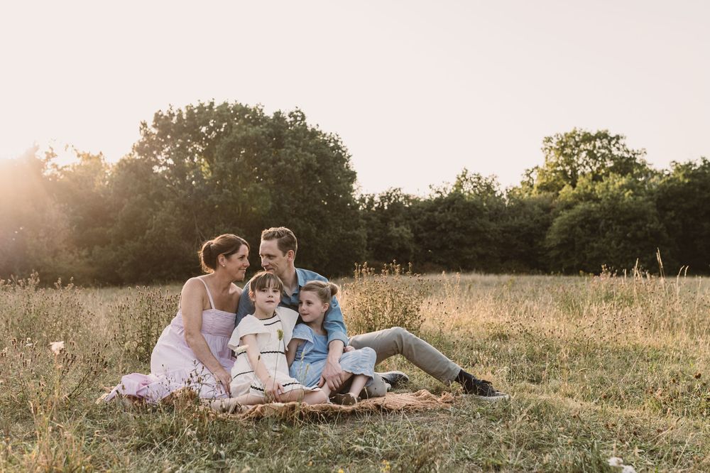 Family of four sitting together on a blanket in a sunny meadow, sharing a warm golden hour moment outdoors.