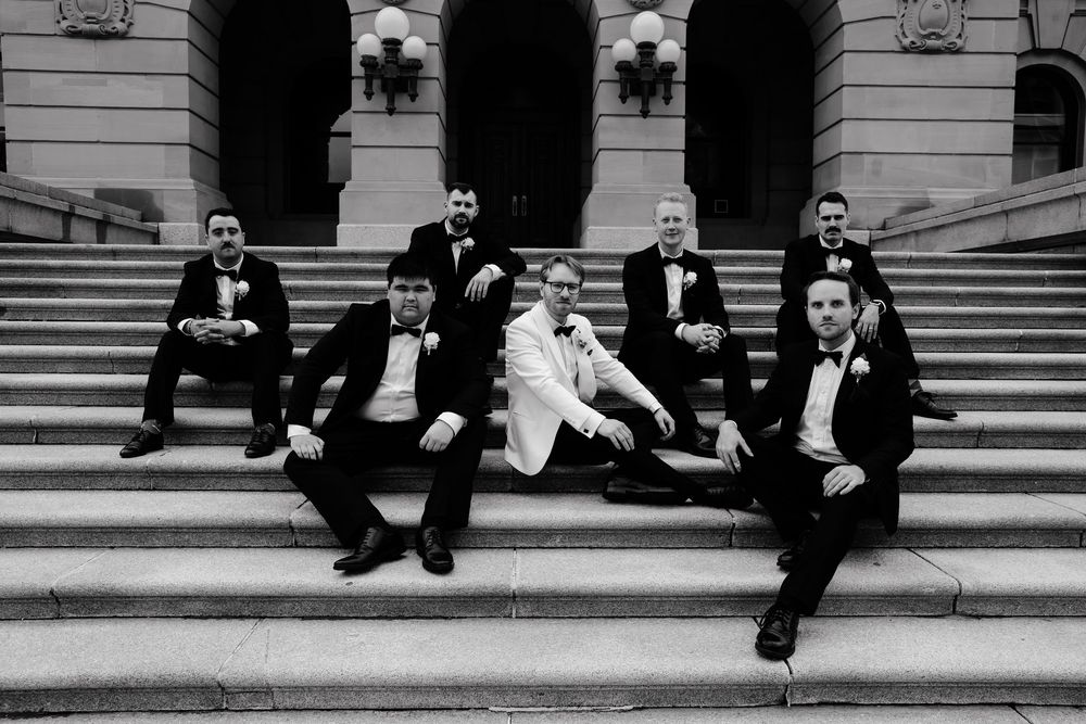 Groom in white tuxedo poses with groomsmen in black suits on stone steps in classic black and white wedding photo.