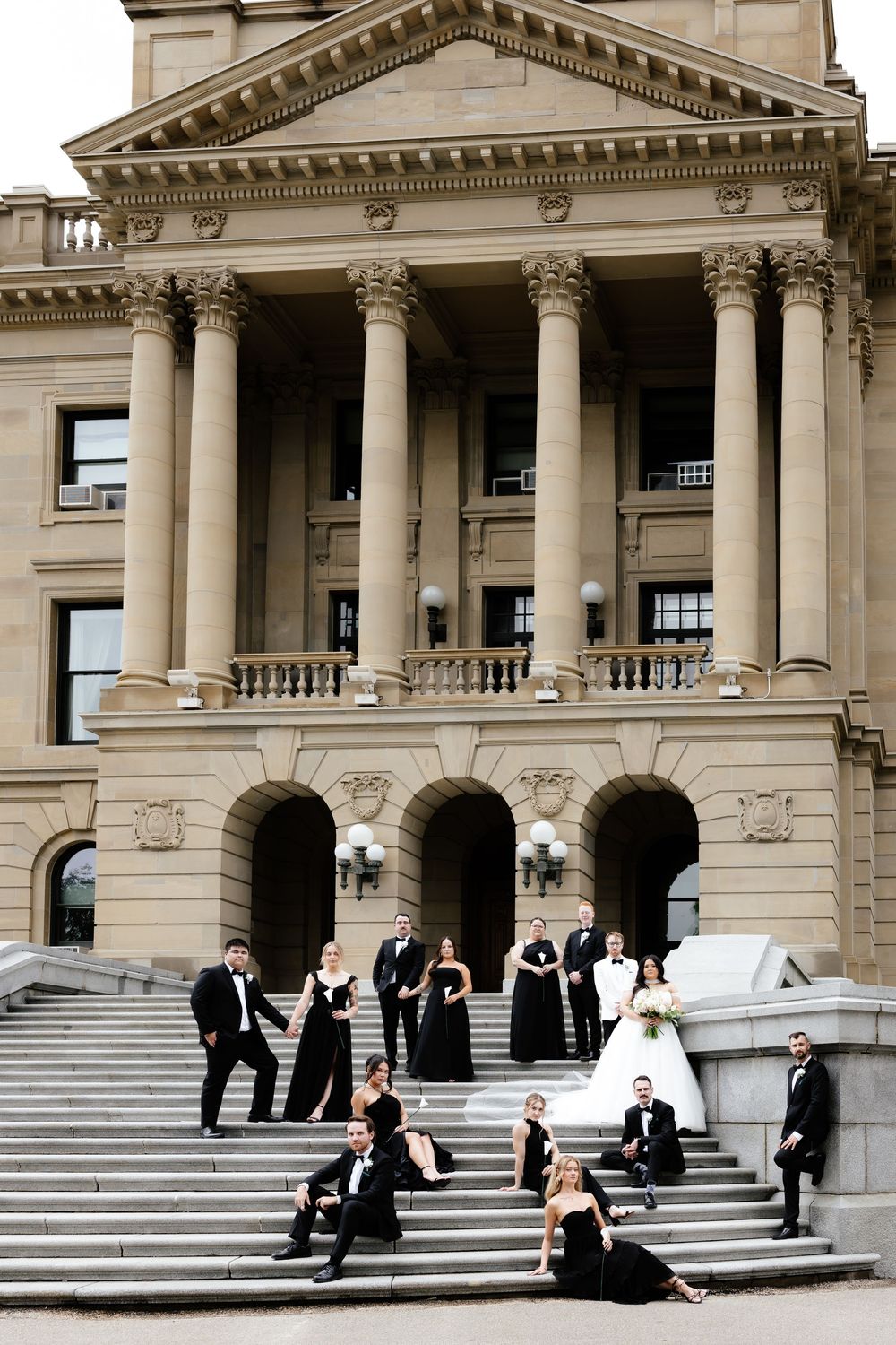Wedding party in black and white attire posing on grand stone steps of a classic government building with columns.