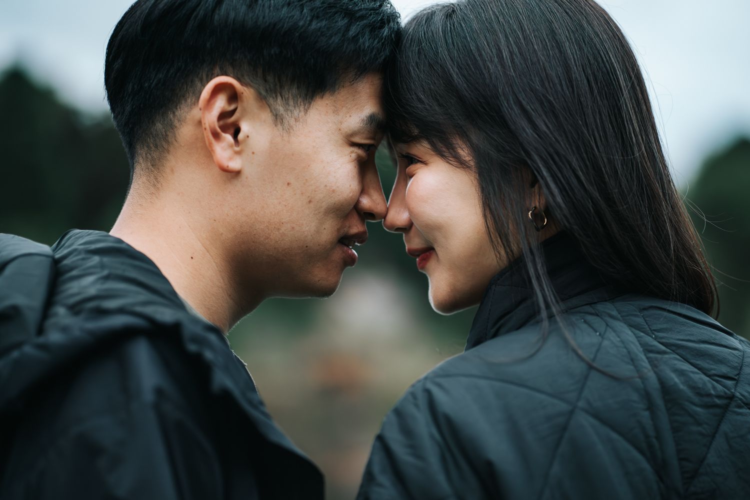 A young couple in black jackets share a romantic forehead-to-forehead moment outdoors taken by Melbourne engagement photographer.