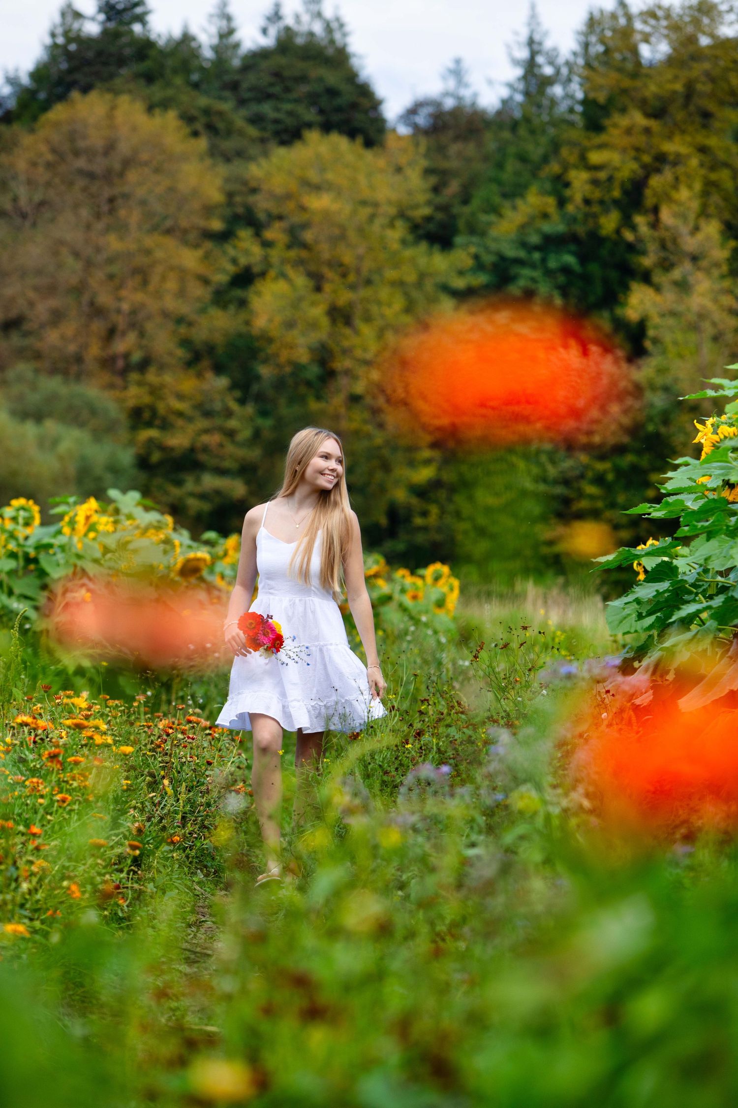 Young blonde woman in white dress walks through vibrant garden holding red flowers, surrounded by sunflowers and colorful blooms.