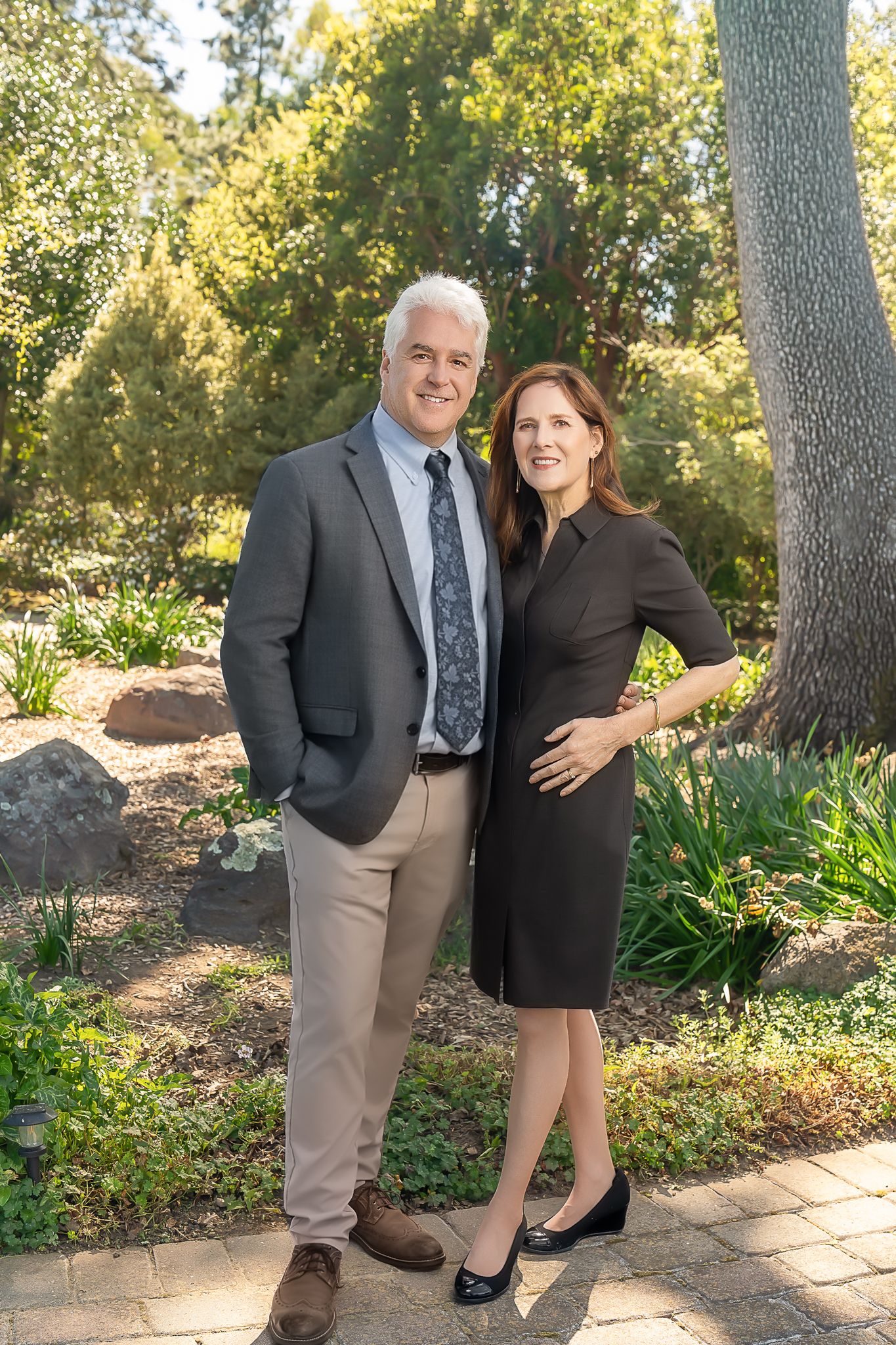 A smiling couple poses together in a lush garden, dressed in professional attire for a formal portrait.