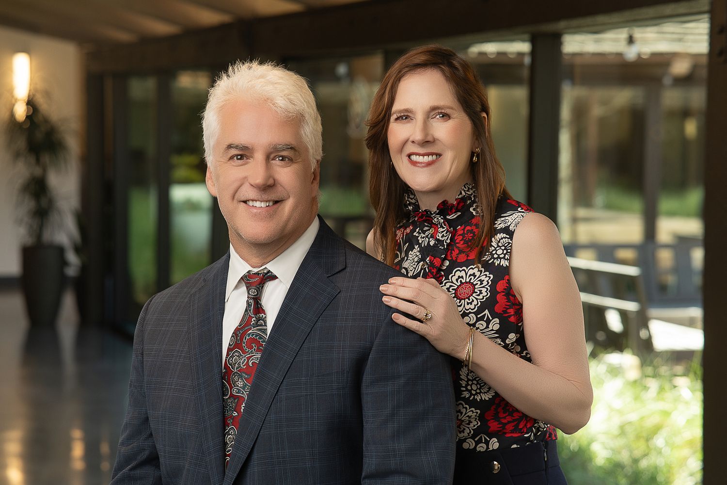 A smiling man in a gray suit and red tie poses with a woman in a floral dress in an elegant indoor setting.