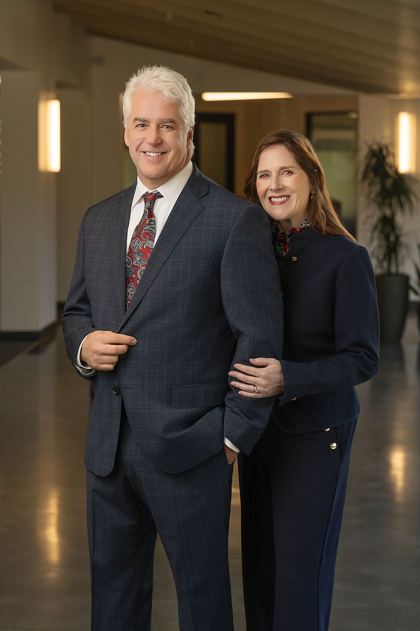 Two professionally dressed adults smiling in a modern office hallway, wearing navy and gray business attire.