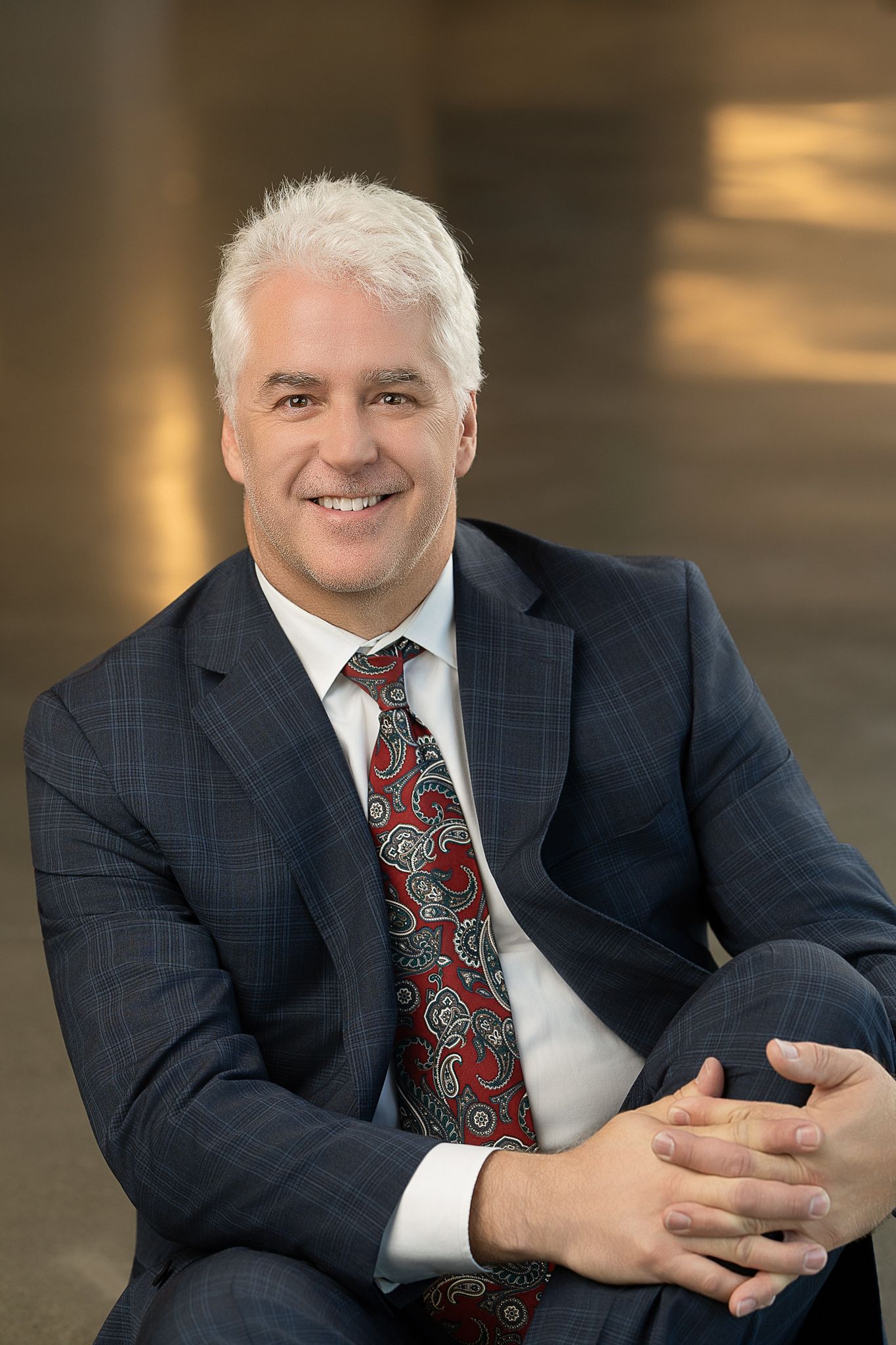 Professional man in navy suit and red paisley tie smiling confidently in a modern corporate setting.