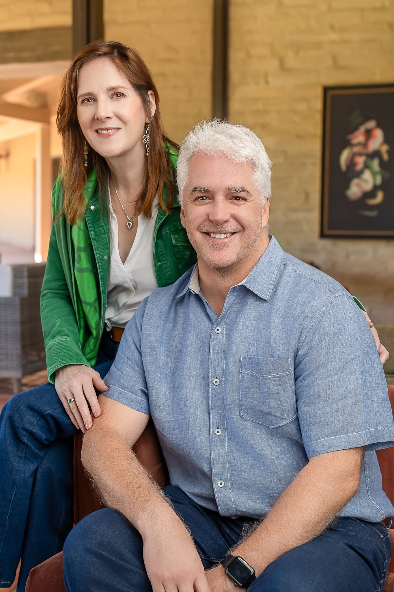 A smiling man and woman pose together indoors, the woman in green jacket standing behind the seated man.
