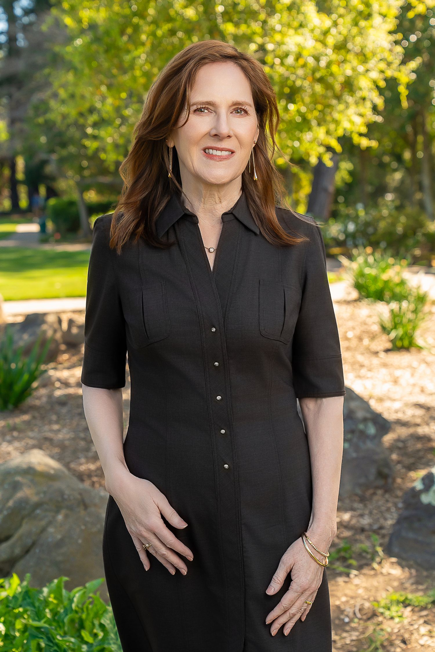 A smiling woman with auburn hair wearing a black dress stands in a sunlit garden with green trees.