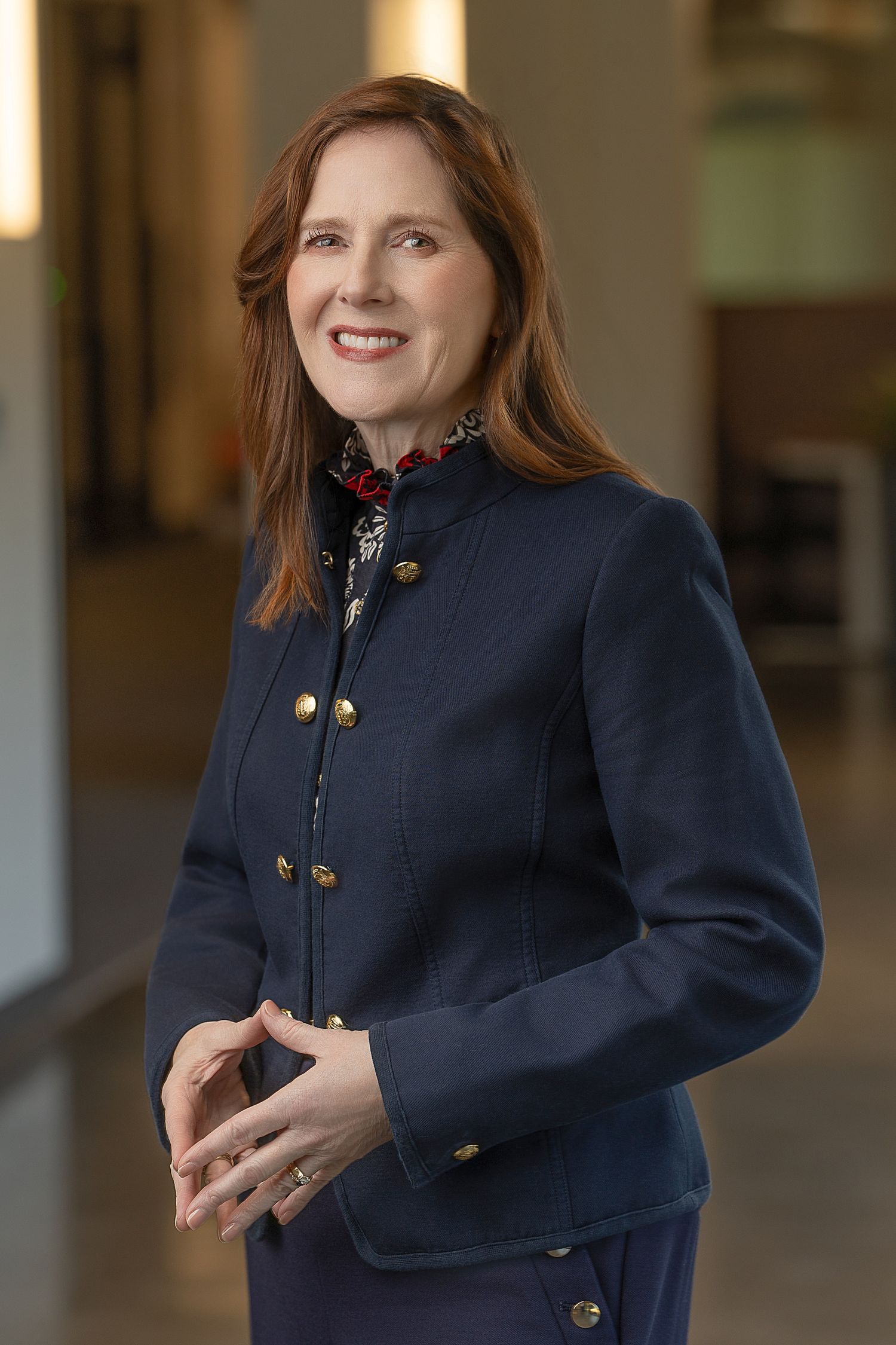 Professional woman in navy blue military-style jacket with gold buttons, smiling in a modern office hallway.