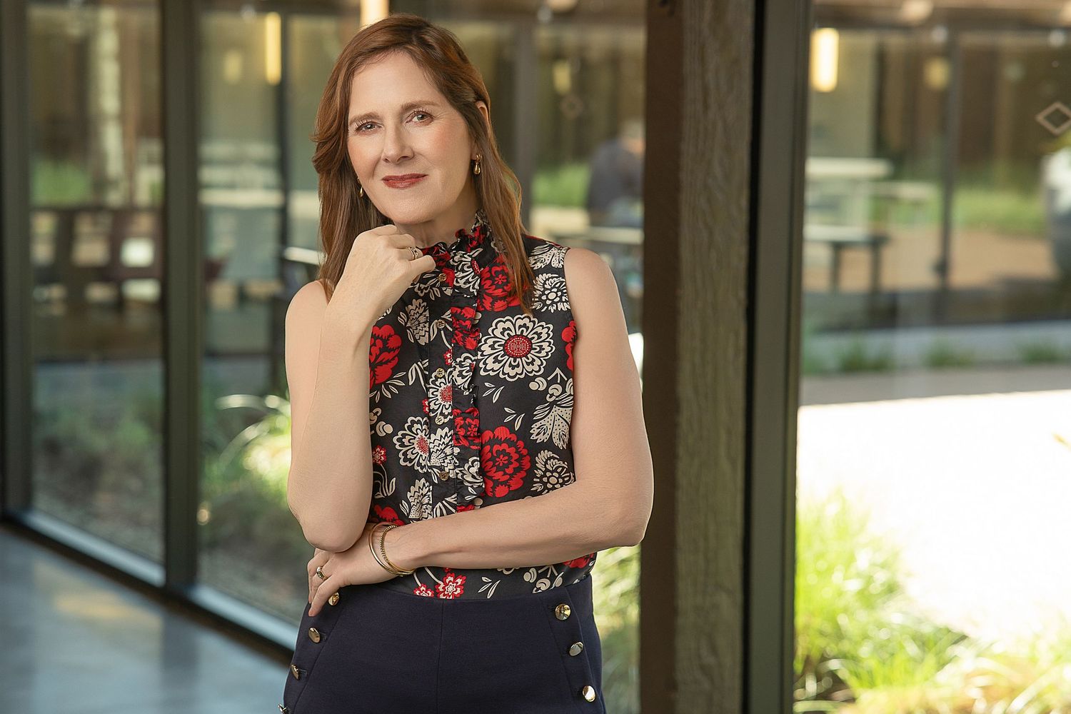Professional woman in floral sleeveless top posing confidently indoors near large windows.