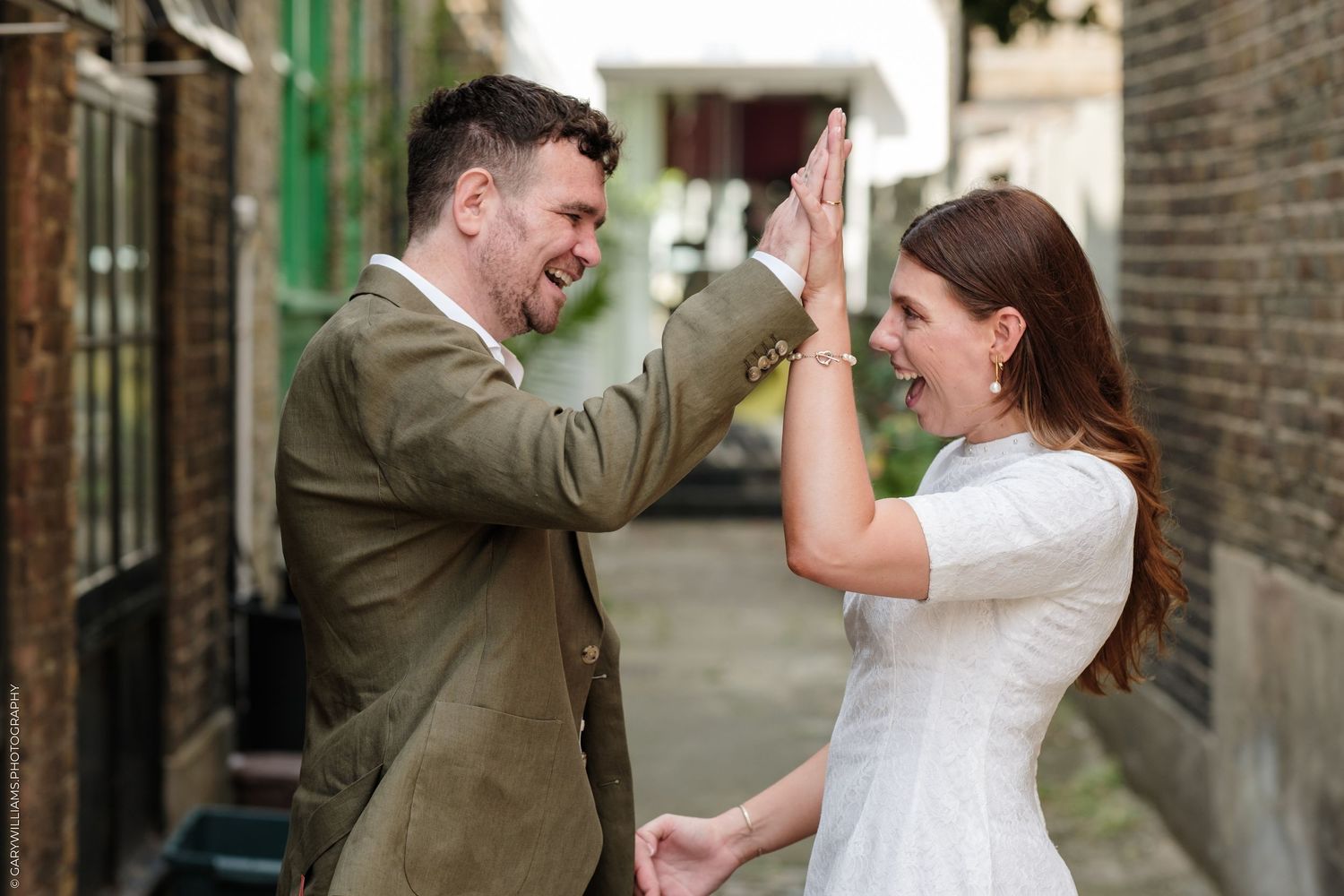Joyful couple in wedding attire high-fiving outdoors in an alley, celebrating their special day.