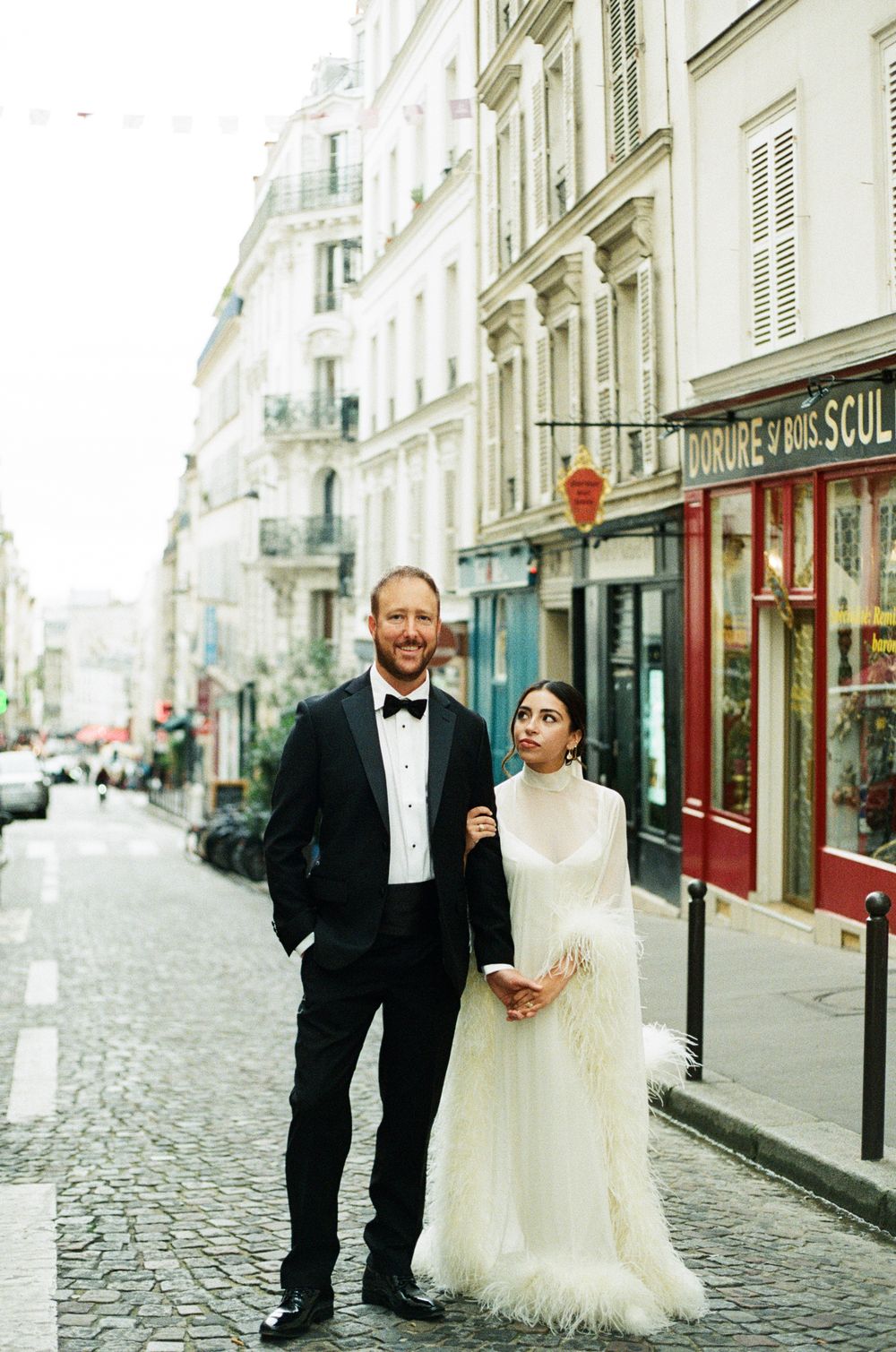 Couple in wedding attire posing on a charming Parisian cobblestone street with classic French architecture.