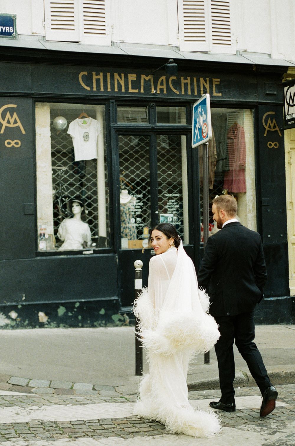 Bride in white feather gown and groom in black tuxedo walking past Chinemachine boutique on a Paris cobblestone street.