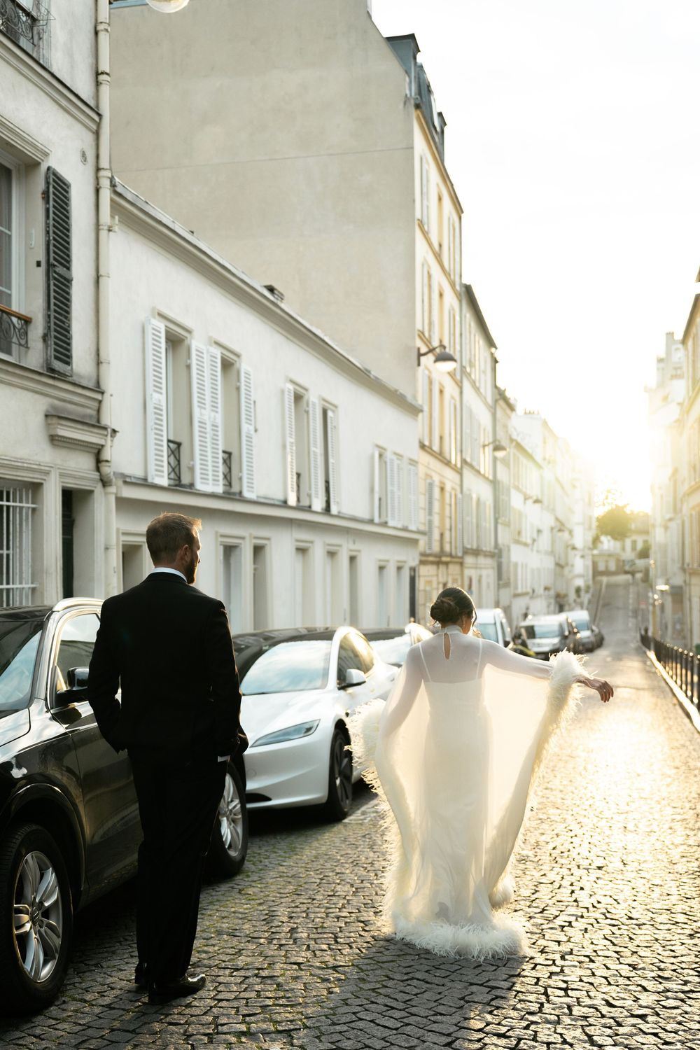 Bride in white gown and groom in black tuxedo walk cobblestone Paris street at golden hour sunset.
