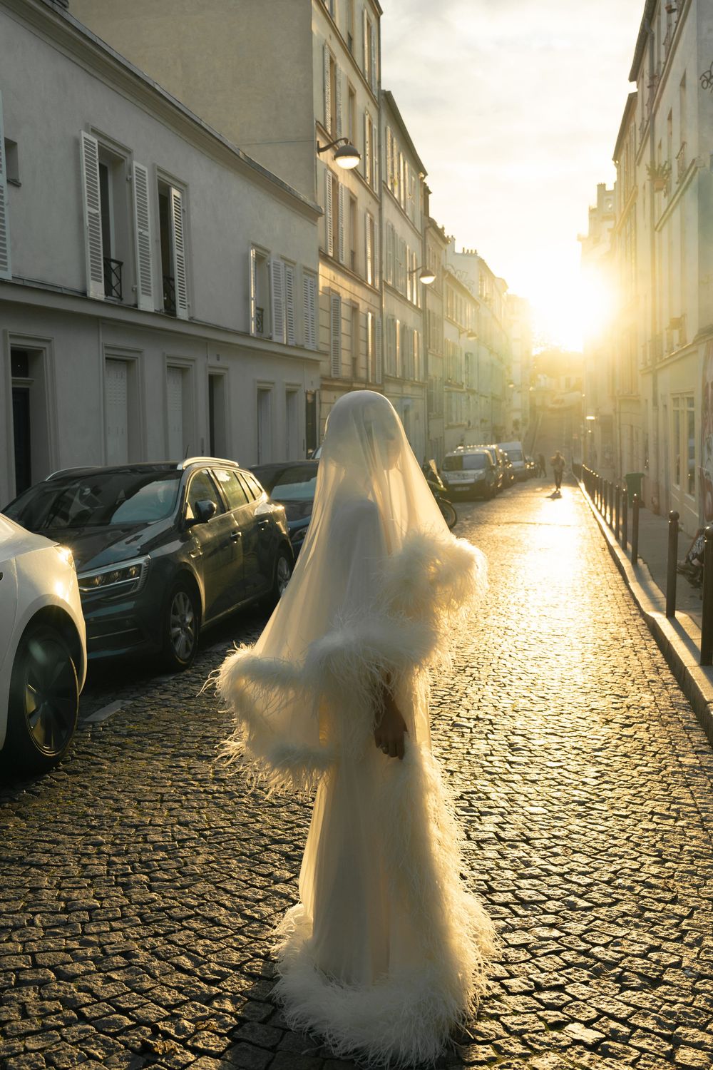 A bride in a flowing white gown and veil walks down a sunlit cobblestone street in Paris at golden hour.