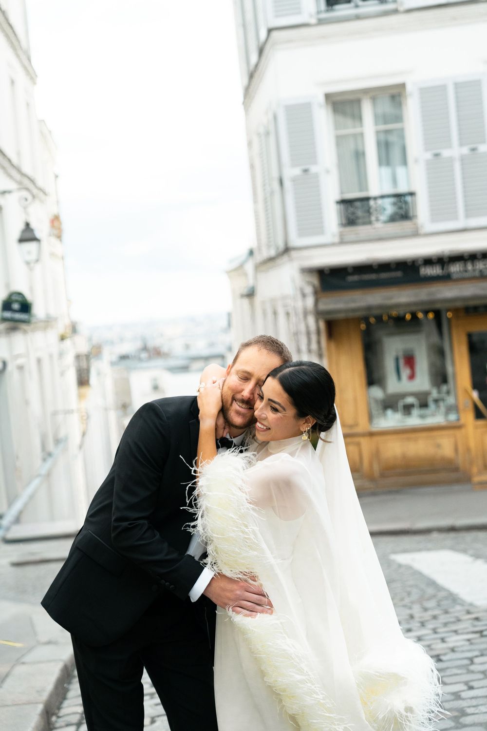 A joyful couple in wedding attire embraces on a cobblestone street in Paris, with charming buildings in the background.