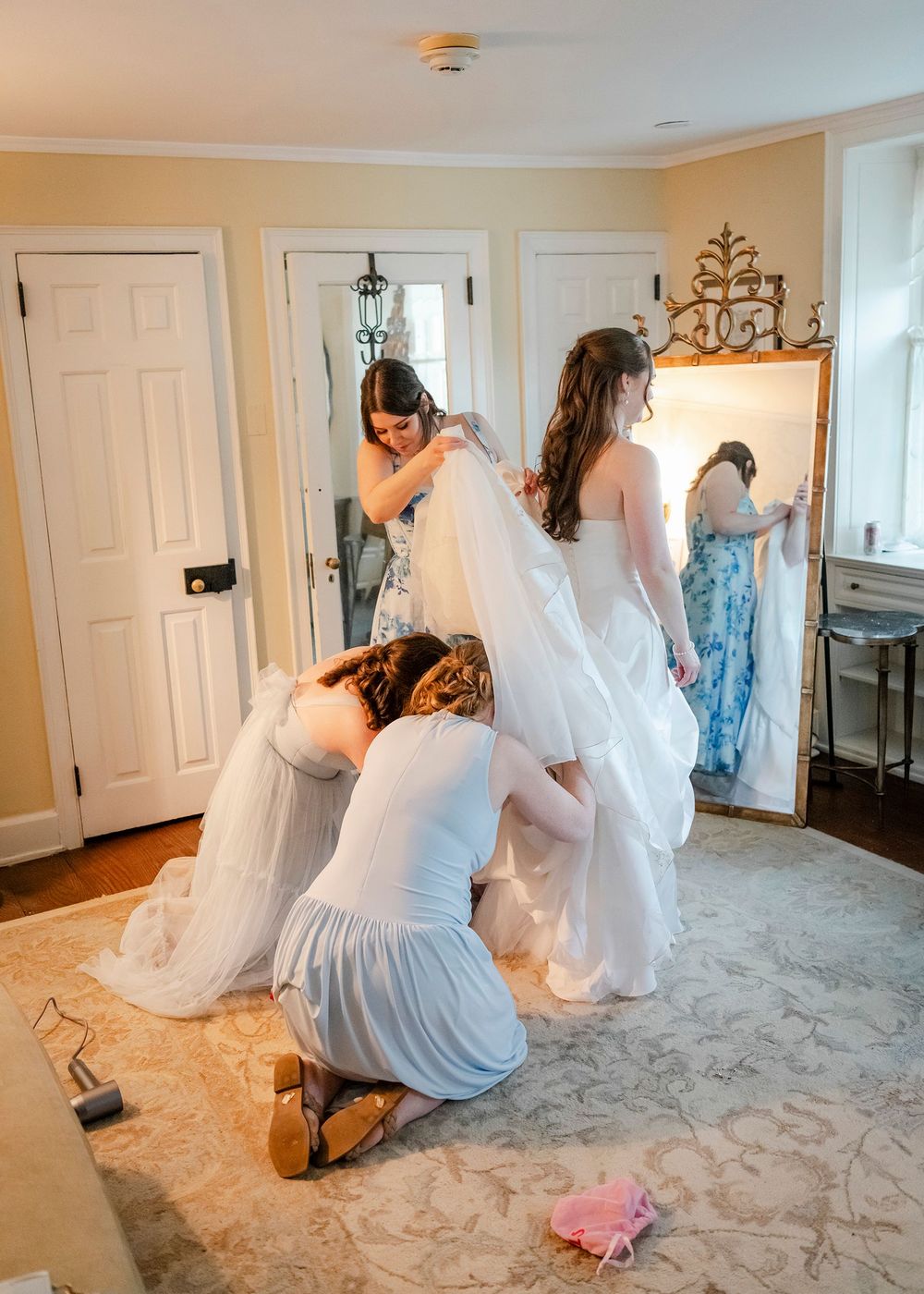 Bridesmaids helping bride into white wedding gown and veil in elegant room with mirror.