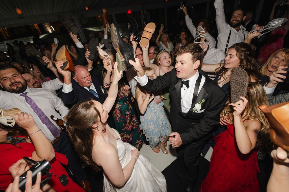 Bride and groom celebrate joyfully on a crowded wedding dance floor with guests raising shoes in the air.