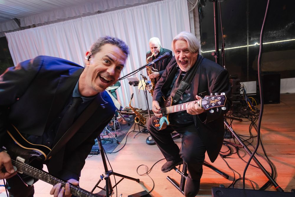 Two guitarists smiling and performing live on stage at an indoor music event with a drummer in the background.