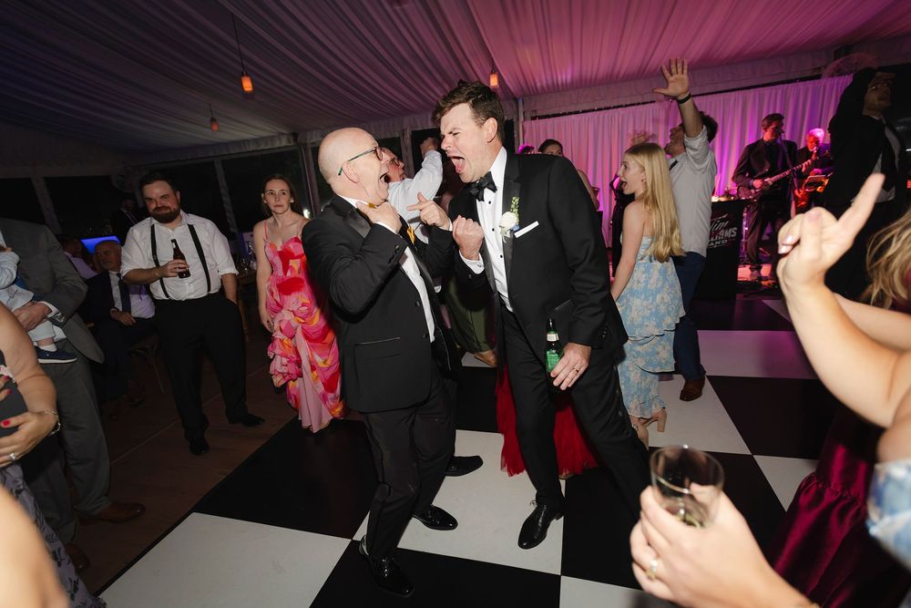 Two men in tuxedos dancing together at a wedding reception on a black and white checkered dance floor.