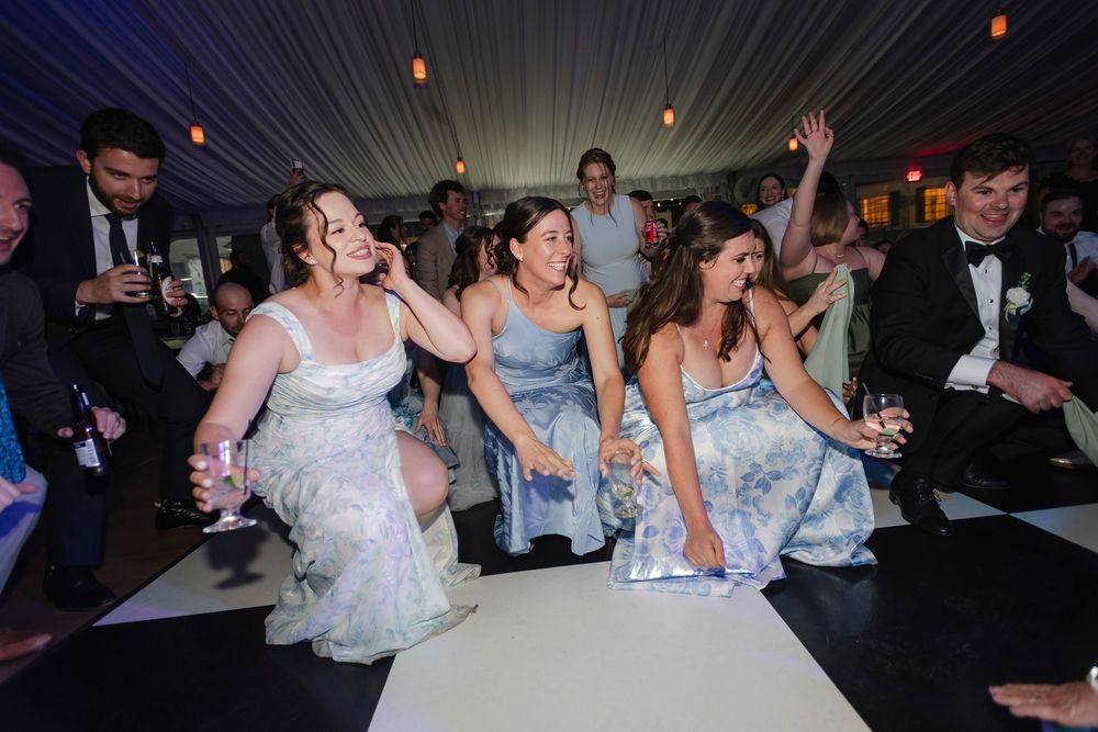 Bridesmaids in floral dresses laugh and dance joyfully at a wedding reception on a white dance floor.