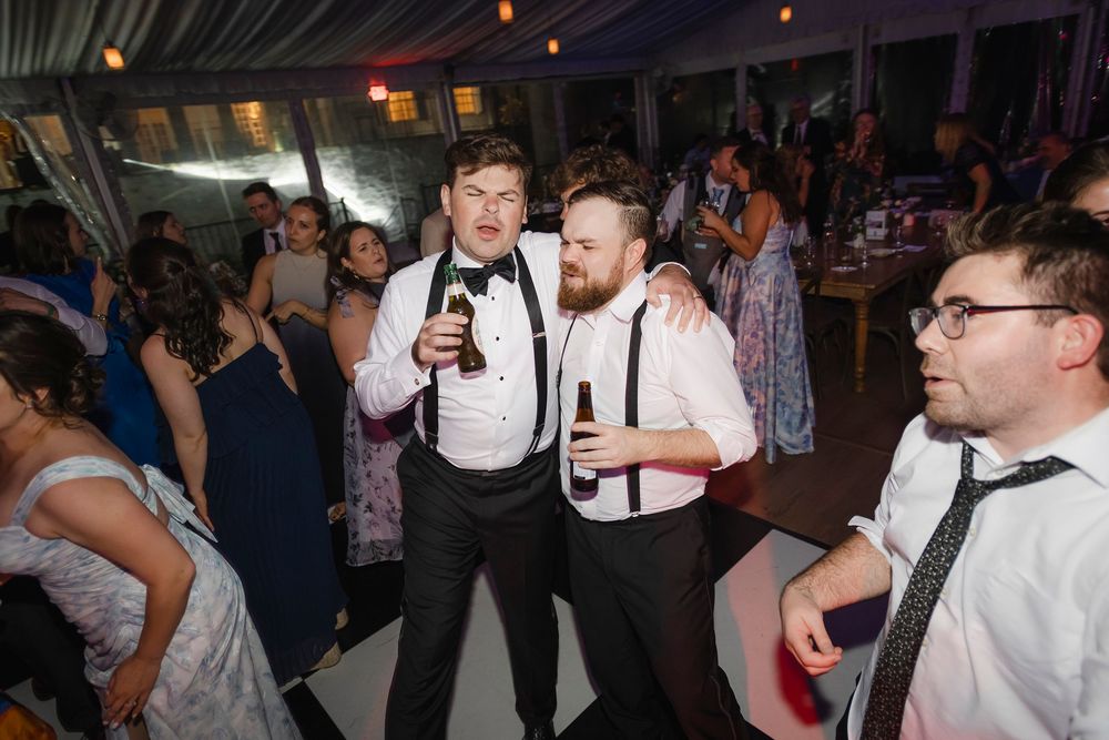 Two men in suspenders and bow ties celebrate with beers on a wedding reception dance floor.